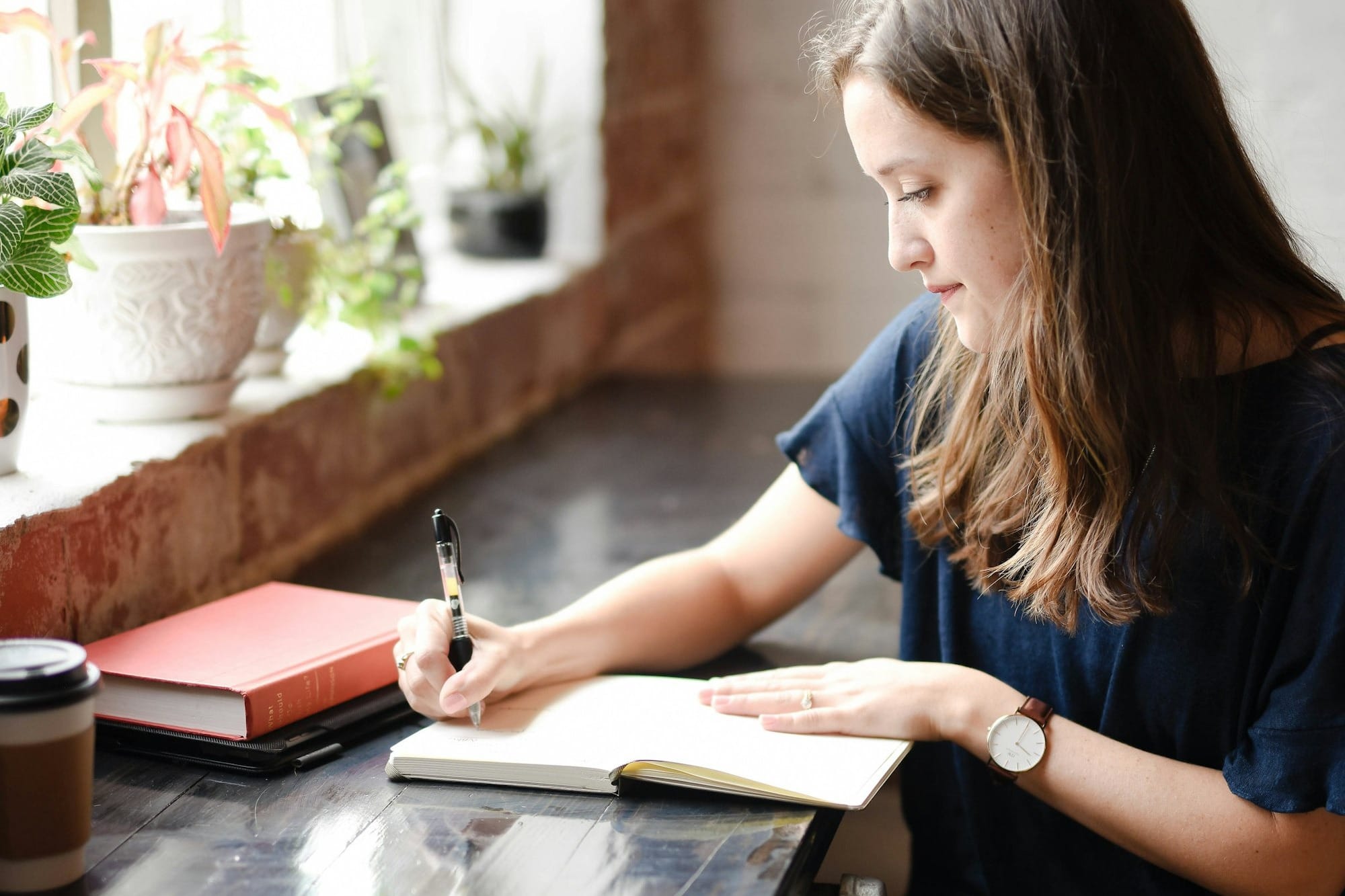 woman sitting in front of black table writing on white book near window